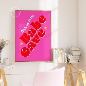 Puede incluir: Un p&oacute;ster rosa y rojo con el texto "Welcome to the Babe Cave" en una fuente retro. El p&oacute;ster est&aacute; enmarcado en oro y tiene dos estrellas amarillas.