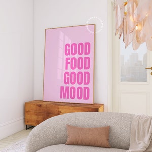 May include: A pink framed poster with the words "GOOD FOOD GOOD MOOD" in bold pink letters. The poster is displayed on a wooden cabinet in a room with a neutral colour palette. A beige sofa and a brown cushion are in the foreground.