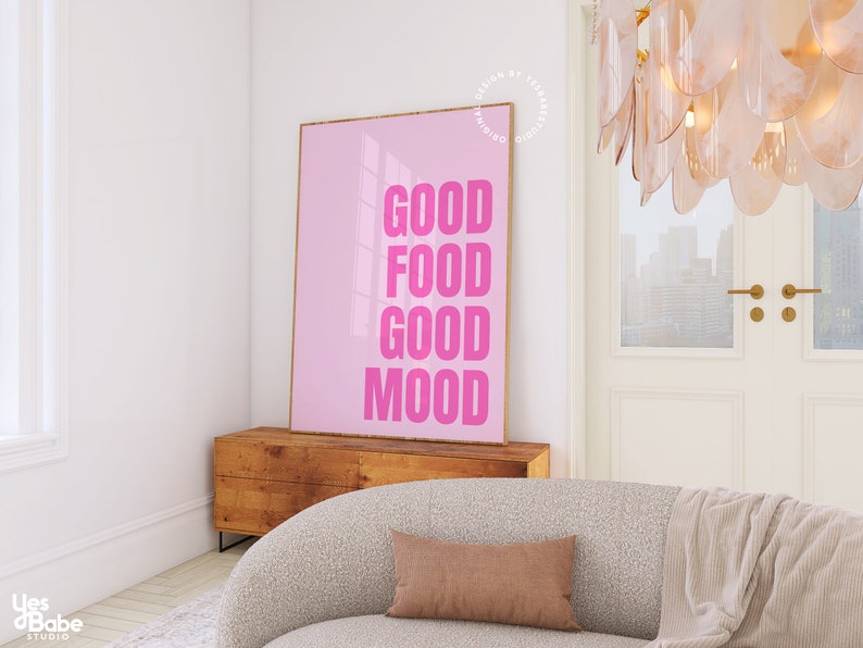 May include: A pink framed poster with the words "GOOD FOOD GOOD MOOD" in bold pink letters. The poster is displayed on a wooden cabinet in a room with a neutral colour palette. A beige sofa and a brown cushion are in the foreground.