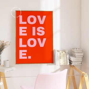 Może przedstawiać: Wydruk w ramie z czerwonym tłem i tekstem "LOVE IS LOVE." w r&oacute;żowych literach. Wydruk wisi na białej ścianie nad drewnianym biurkiem z r&oacute;żowym krzesłem i wazą z kwiatami.