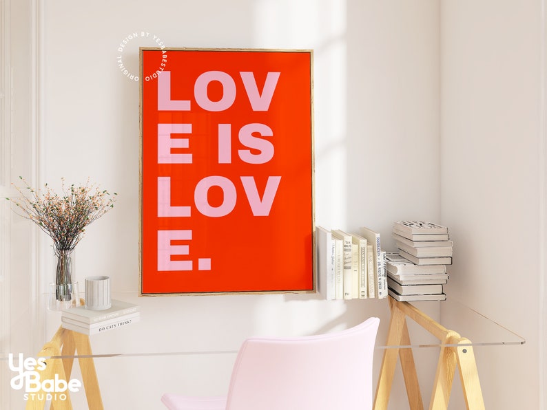 Może przedstawiać: Wydruk w ramie z czerwonym tłem i tekstem "LOVE IS LOVE." w r&oacute;żowych literach. Wydruk wisi na białej ścianie nad drewnianym biurkiem z r&oacute;żowym krzesłem i wazą z kwiatami.