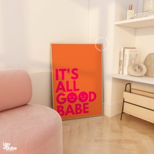 May include: An orange framed art print with the words "IT'S ALL GOOD BABE" in bright pink. The print features two smiley face emojis. The art is displayed next to a pink velvet chair in a well-lit room.