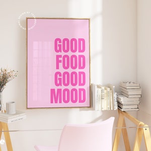 May include: A pink framed print with the words "GOOD FOOD GOOD MOOD" in bright pink. The art is displayed in a room with a clear desk, a pink chair, and a small table with books and a vase of flowers.