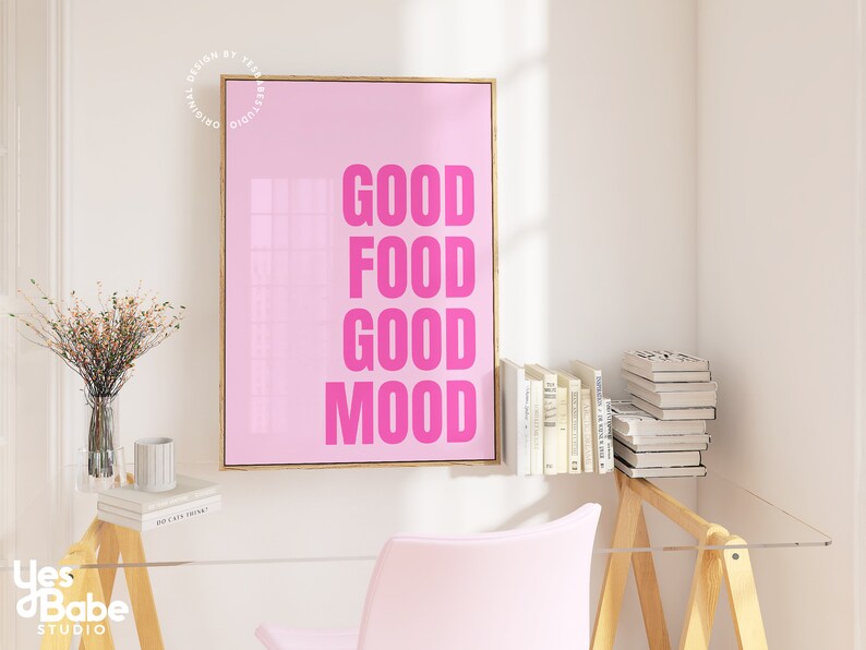 May include: A pink framed print with the words "GOOD FOOD GOOD MOOD" in bright pink. The art is displayed in a room with a clear desk, a pink chair, and a small table with books and a vase of flowers.