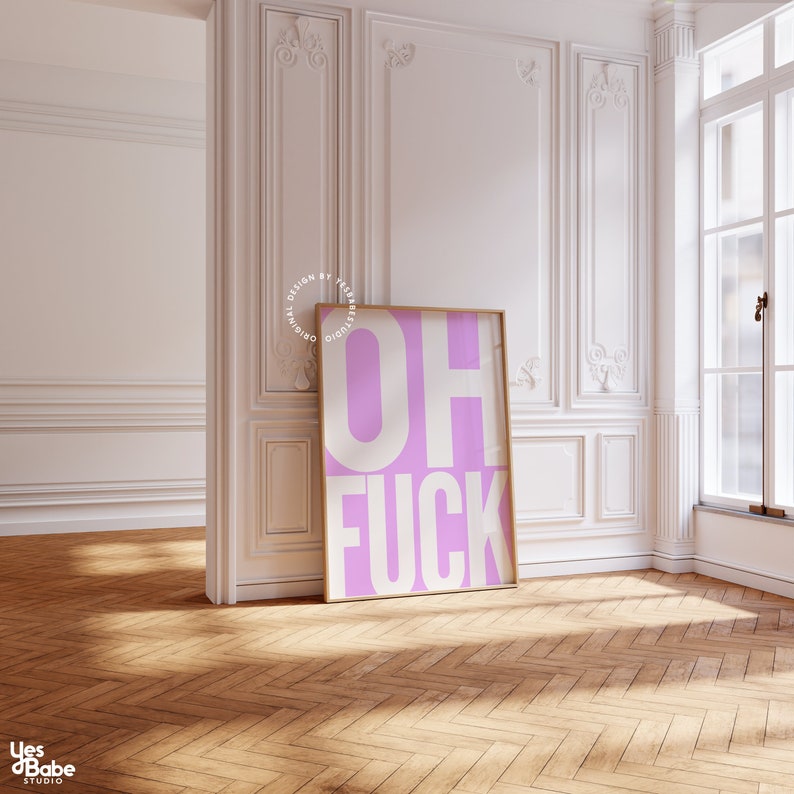 May include: A framed print with a pink background and white text that reads "OH FUCK". The print is hanging on a white wall in a room with white wainscoting and hardwood floors.