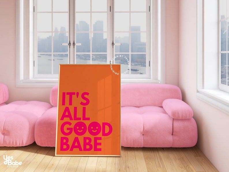 May include: An orange art print with the phrase "IT'S ALL GOOD BABE" in bright pink, with two smiley faces. The print is displayed in a gold frame, leaning against a pink sofa in a room with a window.