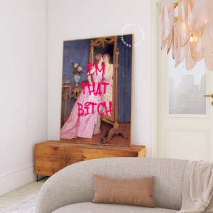 May include: A framed print of a vintage painting of two women in a room with a mirror. The woman in the foreground is wearing a pink dress and the woman in the mirror is wearing a white dress. The text "I'M THAT BITCH" is written in pink letters over the image.