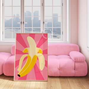 May include: A pink and yellow banana print poster with a retro style design. The poster is framed in a gold frame and is hanging on a wall in a room with a pink sofa.