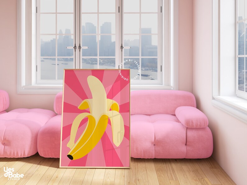 May include: A pink and yellow banana print poster with a retro style design. The poster is framed in a gold frame and is hanging on a wall in a room with a pink sofa.