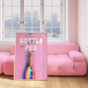 May include: A pink poster with a gold frame featuring a rainbow-colored champagne bottle with the text "Bottle Fed" in white.