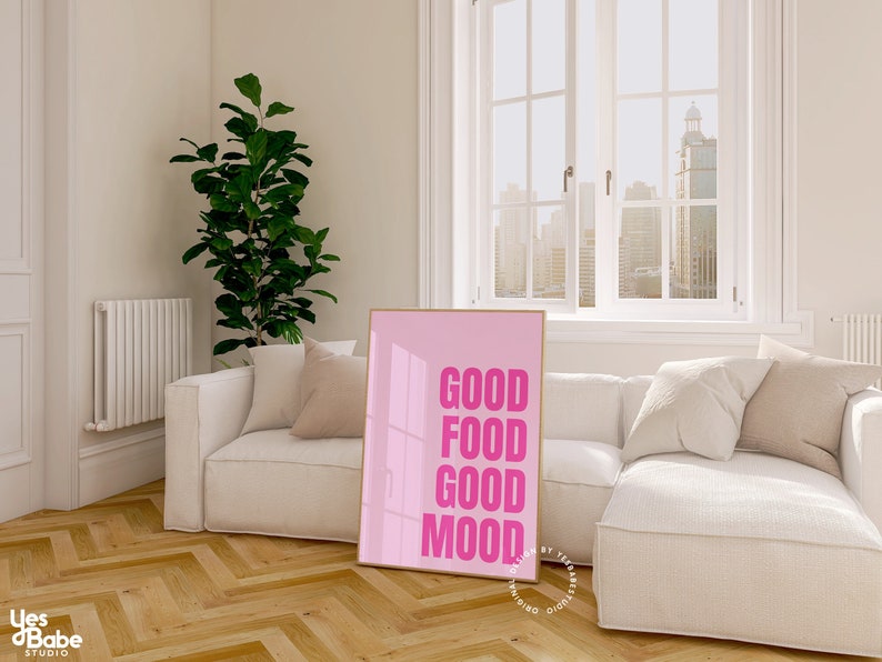 May include: A pink framed print with the words "GOOD FOOD GOOD MOOD" in bold pink letters. The print is in a modern living room with a white sofa, cushions, and a large window overlooking a cityscape.