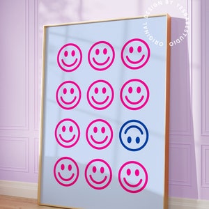 Pu&ograve; includere: Una stampa incorniciata con uno sfondo blu chiaro e una griglia di 12 facce sorridenti rosa. Una faccia sorridente &egrave; blu e capovolta. La stampa ha una cornice dorata e il testo "Original Design by Yes Babe Studio" &egrave; stampato in un cerchio attorno alla cornice.