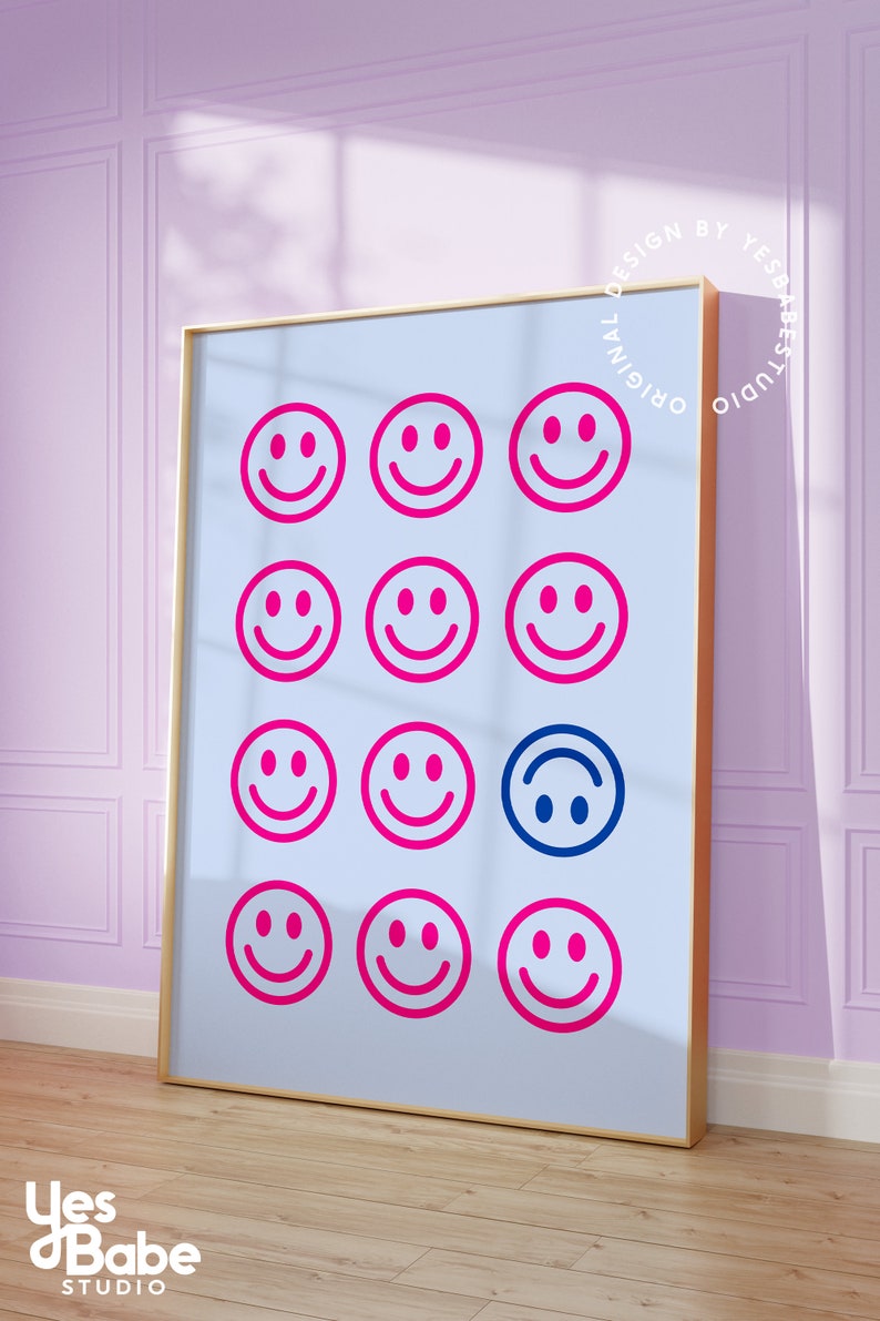 Pu&ograve; includere: Una stampa incorniciata con uno sfondo blu chiaro e una griglia di 12 facce sorridenti rosa. Una faccia sorridente &egrave; blu e capovolta. La stampa ha una cornice dorata e il testo "Original Design by Yes Babe Studio" &egrave; stampato in un cerchio attorno alla cornice.