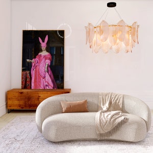 May include: A white curved sofa with a brown throw pillow and a beige throw blanket. The sofa is in a living room with a white wall, a wooden cabinet, and a large painting of a woman wearing a pink dress and a pink bunny mask. A large, round, chandelier with clear glass shades hangs from the ceiling.