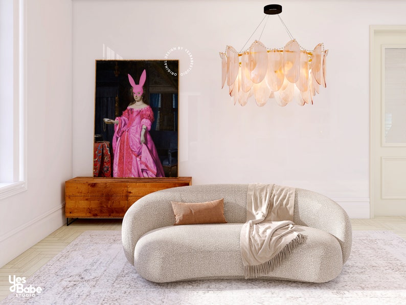 May include: A white curved sofa with a brown throw pillow and a beige throw blanket. The sofa is in a living room with a white wall, a wooden cabinet, and a large painting of a woman wearing a pink dress and a pink bunny mask. A large, round, chandelier with clear glass shades hangs from the ceiling.