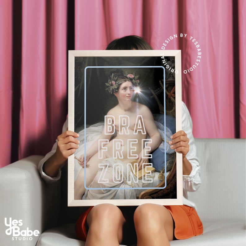 May include: A woman holds a framed print of a painting of a woman with a crown of flowers and a book. The print has the text "BRA FREE ZONE" in neon pink letters.