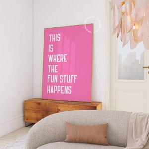 Puede incluir: P&oacute;ster rosa con texto blanco que dice "This is where the fun stuff happens". El p&oacute;ster est&aacute; colgado en una pared blanca en una habitaci&oacute;n con un sof&aacute; y una c&oacute;moda de madera.