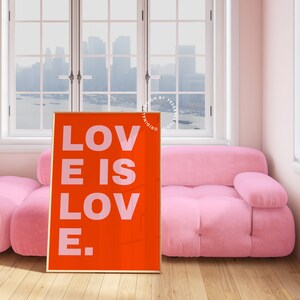Może przedstawiać: Wydruk w ramie z czerwonym tłem i białym tekstem, kt&oacute;ry brzmi "LOVE IS LOVE." Wydruk wisi na ścianie w pokoju z r&oacute;żową sofą i oknem z widokiem na panoramę miasta.