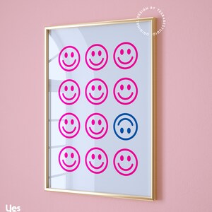 Pu&ograve; includere: Una stampa incorniciata con sfondo bianco e una griglia di 12 facce sorridenti rosa. La faccia sorridente al centro in basso &egrave; blu e capovolta. La cornice &egrave; dorata.