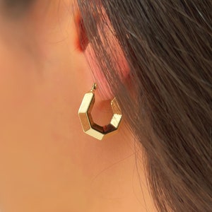 Op de afbeelding: Een close-up van een goudkleurige geometrische oorring. De oorring heeft een facetontwerp, met meerdere platte oppervlakken die een polygonale vorm creëren. De oorring wordt gedragen aan het oor van een persoon met donker haar.