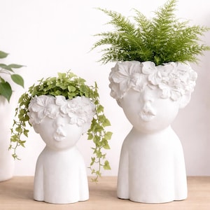 Könnte beinhalten: Zwei weiße Keramik-Pflanzgefäße in Form stilisierter menschlicher Figuren. Die Pflanzgefäße haben eine matte Oberfläche und zeigen ein Gesicht mit einem Blumenkranz. Ein Pflanzgefäß ist kleiner als das andere. Grüne Pflanzen wachsen aus der Oberseite jedes Pflanzgefäßes.