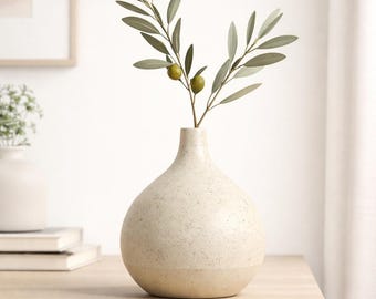 May include: A beige ceramic vase with a speckled texture holds an olive branch with green olives. The vase has a rounded body and a narrow neck. The olive branch has green leaves and two olives. The vase is on a light wood surface.