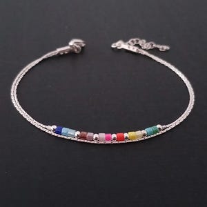 Può includere: Un delicato braccialetto d'argento con una fila di piccole perline colorate. Le perline sono disposte in un motivo arcobaleno, con colori tra cui blu, verde, giallo e rosa. Il braccialetto ha un design semplice ed elegante.