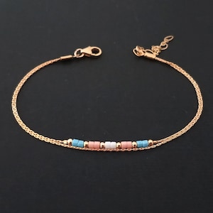 May include: Delicate gold-toned bracelet with a double chain design. The bracelet features a series of small, colorful beads in light blue, pink, and white. The bracelet has a lobster clasp closure and an adjustable chain for a comfortable fit.