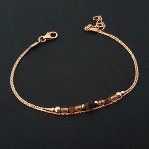Pode incluir: Pulseira em ouro rosa com um design de corrente delicada. A pulseira é adornada com uma série de pequenas contas redondas em tons de castanho e dourado. Um fecho lagosta prende a pulseira. A joia é exibida sobre um fundo escuro.