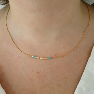 Op de afbeelding: Een delicate gouden ketting met kleine, kleurrijke kralen. De kralen zijn turquoise, perzik en wit, geregen op een fijne gouden ketting. De ketting wordt getoond tegen een huidskleurige achtergrond.