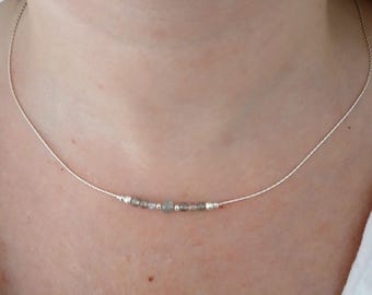 May include: A delicate silver necklace featuring small, round, gray beads. The beads are evenly spaced along a thin, silver chain. The necklace is worn around the neck, showcasing a minimalist jewelry design.