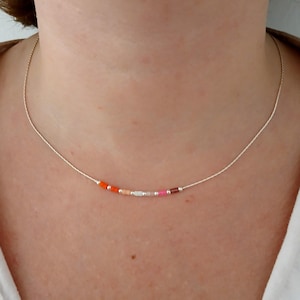 Puede incluir: Un delicado collar con una fina cadena plateada adornada con una serie de pequeñas cuentas de colores. Las cuentas están dispuestas en una fila, mostrando tonos de naranja, rosa y blanco. El collar está diseñado para usarse alrededor del cuello.