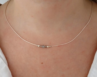 May include: Delicate silver necklace featuring small, light-colored beads. The necklace is strung on a thin, silver chain and rests on a person's neck. The beads are spaced evenly along the chain, creating a minimalist and elegant design.