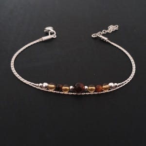 Può includere: Un bracciale in argento con un design a catena delicata. Il bracciale presenta una fila di perline di pietre preziose ambra e marroni. Il bracciale è esposto su una superficie nera.