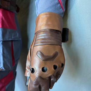Puede incluir: Guantes de cuero marr&oacute;n con agujeros para los dedos y una correa de cuero marr&oacute;n en la mu&ntilde;eca. Los guantes tienen un dise&ntilde;o texturizado y est&aacute;n hechos de un material duradero.