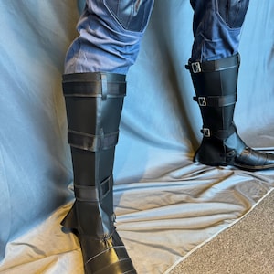Puede incluir: Un par de botas de cuero negro con hebillas y correas. Las botas se llevan sobre pantalones azules con rodilleras.