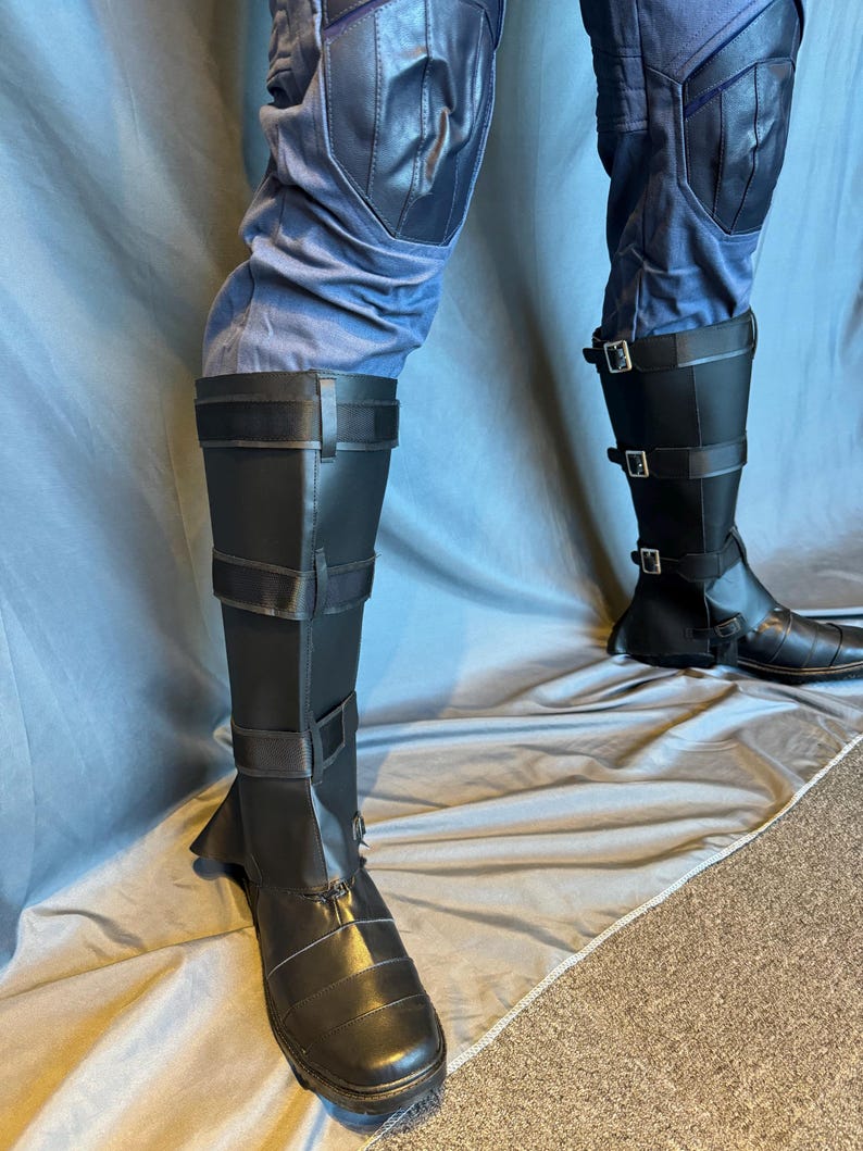 Puede incluir: Un par de botas de cuero negro con hebillas y correas. Las botas se llevan sobre pantalones azules con rodilleras.
