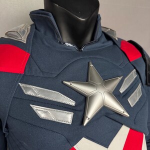 Puede incluir: Un traje de superh&eacute;roe azul y rojo con una estrella plateada en el pecho. El traje tiene un cuello alto y una cremallera en la parte delantera.