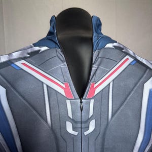 May include: A gray and blue jumpsuit with a detailed geometric design. The suit features red and white accents, a high collar, and a full-length zipper. The fabric has a textured appearance.