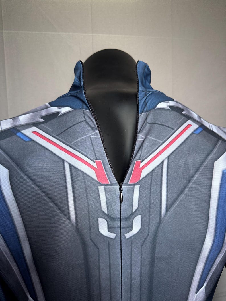 May include: A gray and blue jumpsuit with a detailed geometric design. The suit features red and white accents, a high collar, and a full-length zipper. The fabric has a textured appearance.