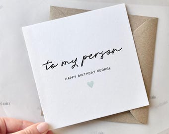 May include: A white greeting card with the text "to my person" in a handwritten font, and "HAPPY BIRTHDAY GEORGE" below. A small, light blue heart is at the bottom. The card is paired with a brown envelope.