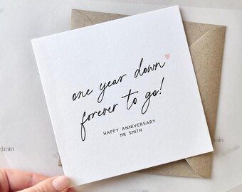May include: A white card with black calligraphy that reads "one year down forever to go!" with a small pink heart. The card also includes the text "Happy Anniversary Mr. Smith".