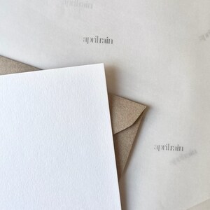 May include: A white card with a textured surface is shown next to a brown envelope. The card is blank and the envelope is closed. The background is a light brown paper with the text "aprilrain" printed on it.