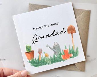 Könnte beinhalten: Eine weiße Geburtstagskarte mit dem Text "Happy Birthday Grandad" in schwarzer Schrift. Die Karte zeigt eine Aquarellillustration von Gartengeräten, einer Topfpflanze und orangefarbenen Gummistiefeln. Sie wird mit einem braunen Umschlag geliefert.
