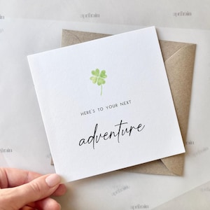 May include: A white card with a green four-leaf clover and the text "Here's to your next adventure".