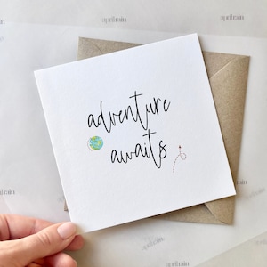 May include: A white card with the text "adventure awaits" in black cursive writing. A small globe and a red arrow are drawn next to the text.
