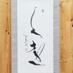 Peut inclure: Peinture sur rouleau japonaise traditionnelle avec calligraphie noire sur papier blanc. Le rouleau est monté sur un tissu gris à motifs et possède des chevilles en bois en haut et en bas. Le fond est un mur en bois clair.