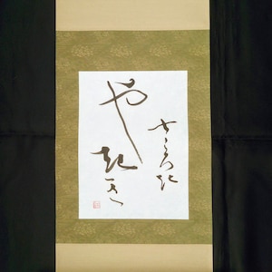 May include: A traditional Japanese scroll with black calligraphy on white paper. The scroll is mounted on a green and gold fabric with a brown wooden dowel at the bottom. The calligraphy reads "Peace".