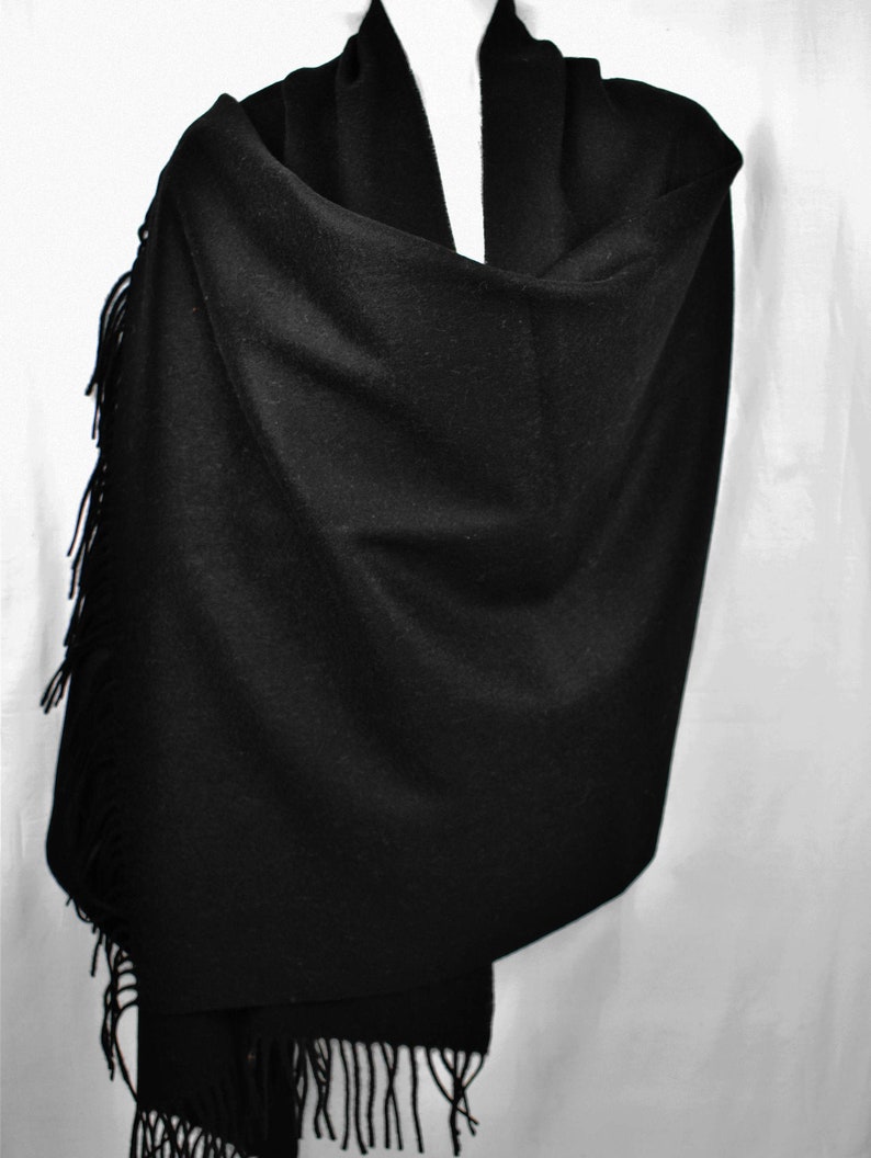 May include: A black fringed shawl made of soft fabric. The shawl is draped over a mannequin and has a simple, elegant design.