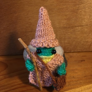 Puede incluir: Un pequeño gnomo de punto con un gorro rosa y una túnica verde, sosteniendo un palo de madera. El gnomo tiene ojos negros y una boca amarilla.